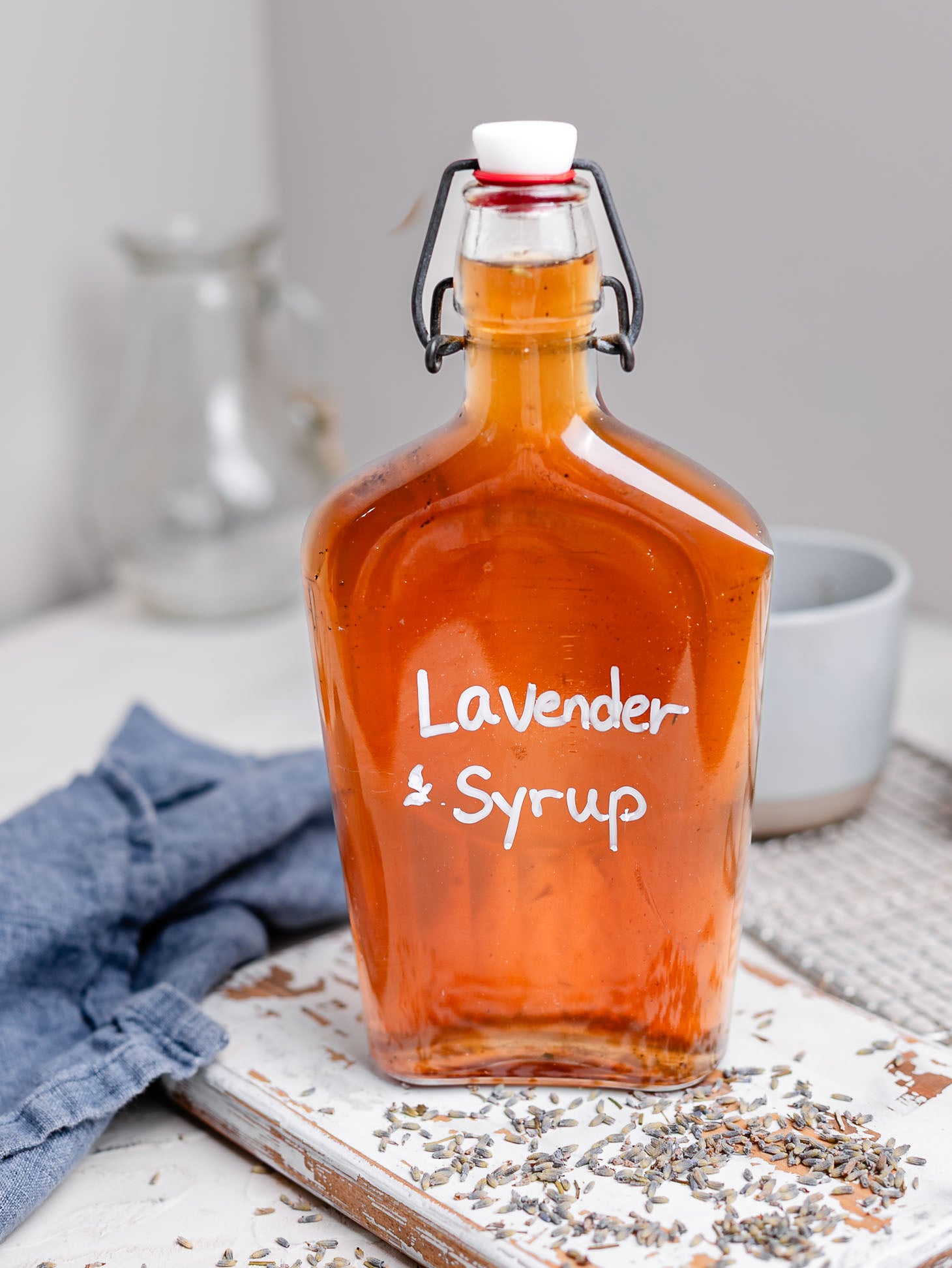 Glass bottle of homemade lavender syrup filled with golden amber syrup and labeled "Lavender Syrup" in white lettering. The bottle sits on a rustic white wooden board scattered with dried lavender buds, alongside a blue linen napkin and coffee mug in the background. A cozy kitchen scene highlighting an easy homemade coffee syrup recipe.
