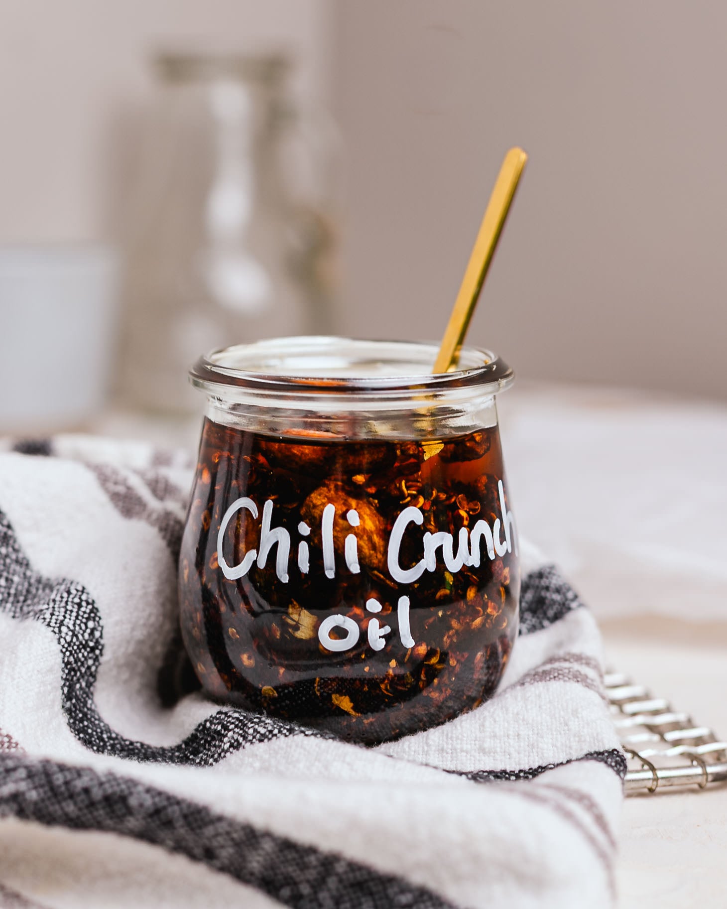Jar of homemade crispy chili oil with chili flakes, crispy garlic, and aromatic spices, photographed on a striped kitchen towel with a gold spoon.