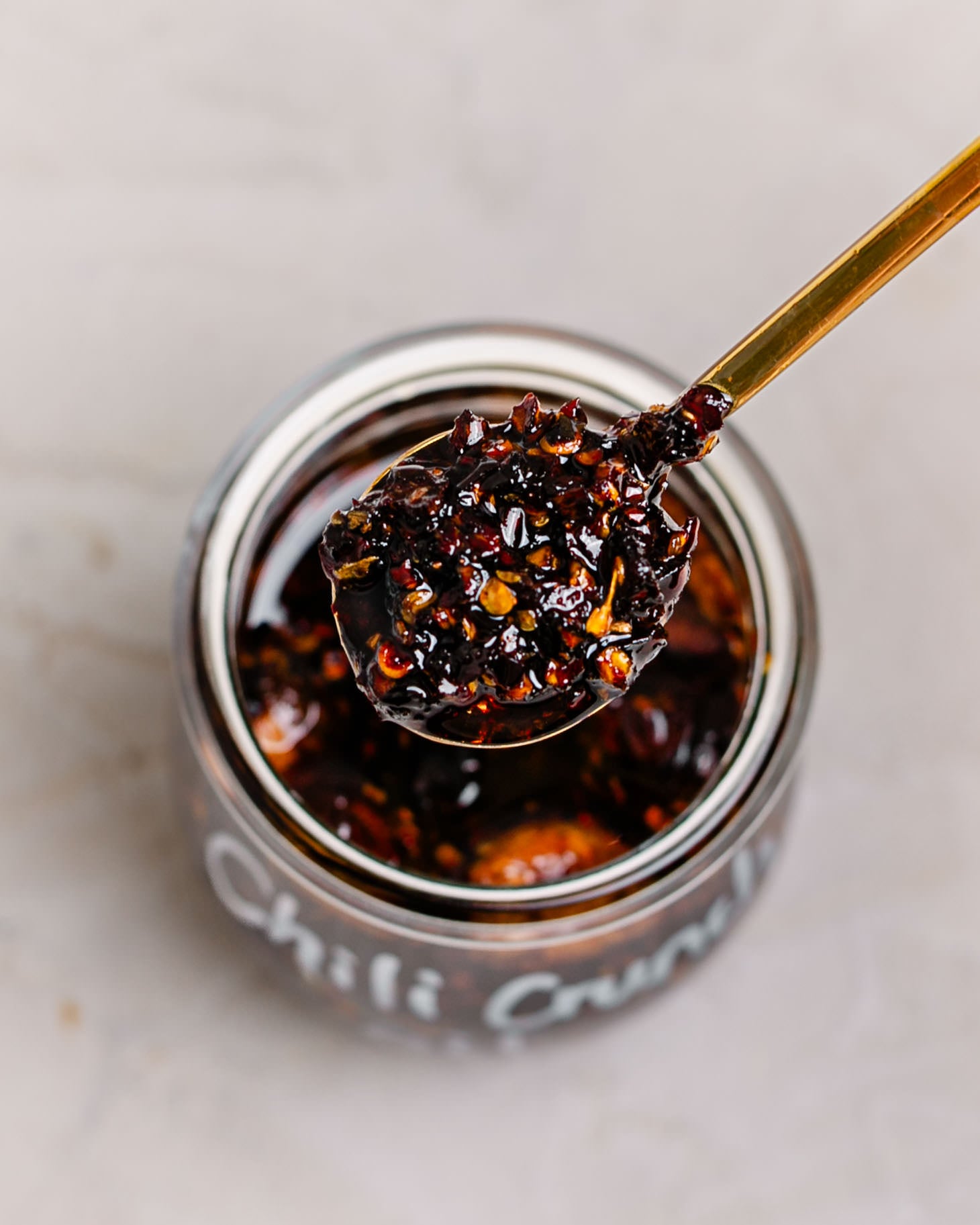 Spoonful of homemade crispy chili oil with crispy garlic, shallots, and chili flakes lifted from a glass jar filled with aromatic chili oil.