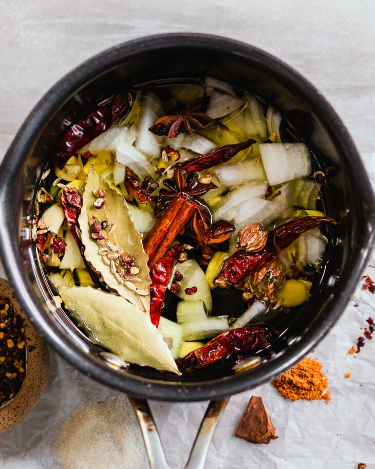 Ingredients for homemade crispy chili oil including garlic, onions, dried chilies, star anise, cinnamon, bay leaves, and Sichuan peppercorns simmering in oil.
