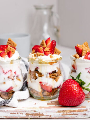 Three strawberry cheesecake Greek yogurt parfaits in glass jars layered with strawberries, vanilla Greek yogurt, graham cracker crumbs, and honey. This easy breakfast meal prep recipe tastes like strawberry cheesecake and is perfect for busy mornings.