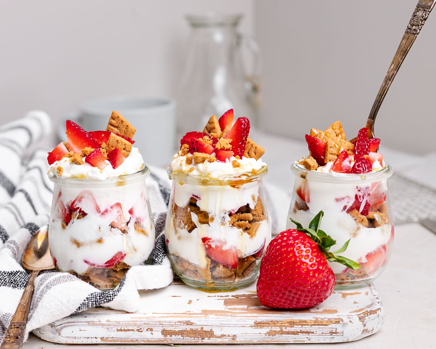Three strawberry cheesecake Greek yogurt parfaits in glass jars layered with strawberries, vanilla Greek yogurt, graham cracker crumbs, and honey. This easy breakfast meal prep recipe tastes like strawberry cheesecake and is perfect for busy mornings.
