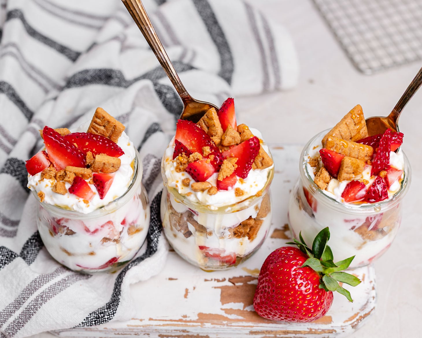 Three strawberry cheesecake Greek yogurt parfaits in glass jars layered with strawberries, vanilla Greek yogurt, graham cracker crumbs, and honey. This easy breakfast meal prep recipe tastes like strawberry cheesecake and is perfect for busy mornings.