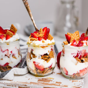 Three strawberry cheesecake Greek yogurt parfaits in glass jars layered with strawberries, vanilla Greek yogurt, graham cracker crumbs, and honey. This easy breakfast meal prep recipe tastes like strawberry cheesecake and is perfect for busy mornings.