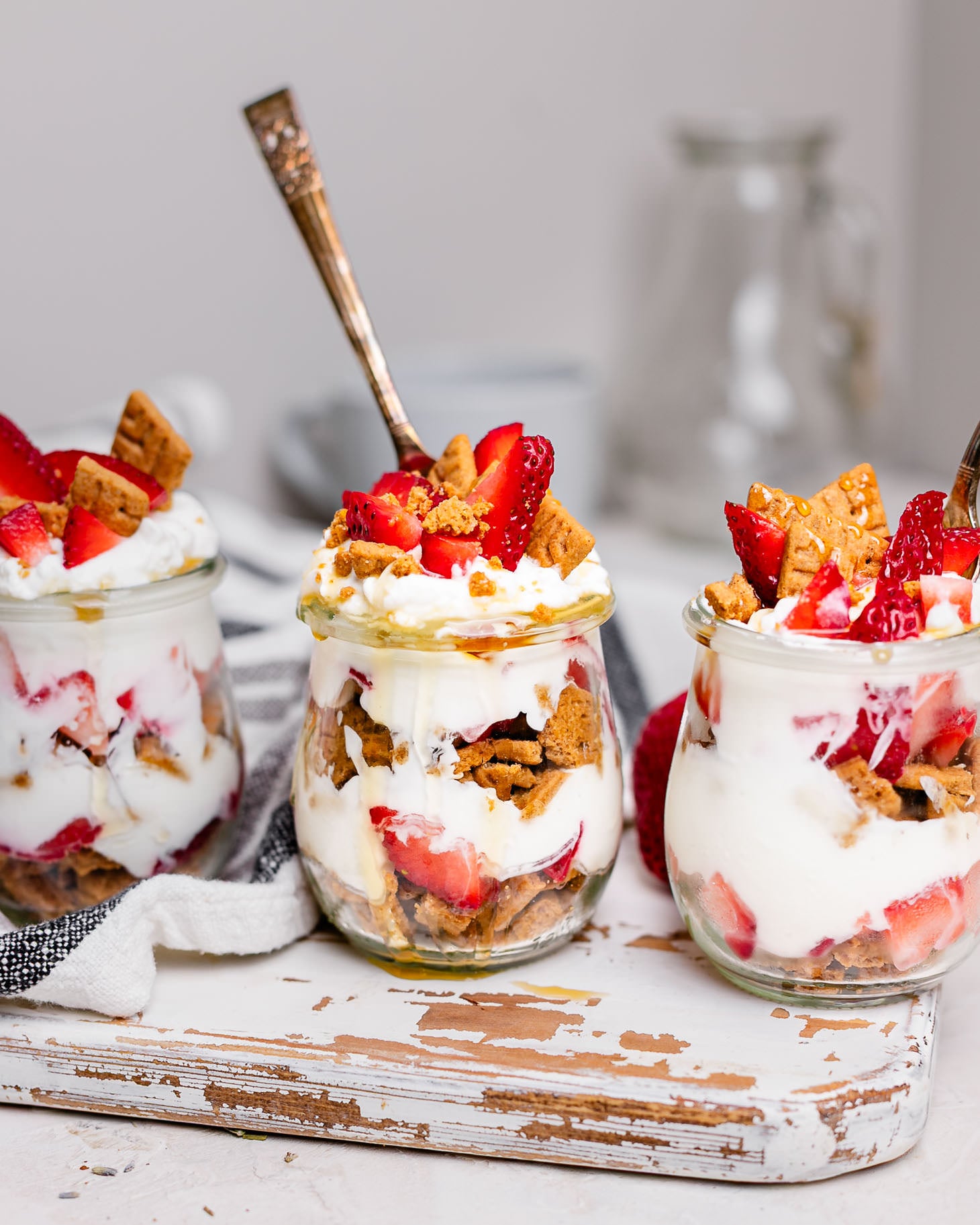 Three strawberry cheesecake Greek yogurt parfaits in glass jars layered with strawberries, vanilla Greek yogurt, graham cracker crumbs, and honey. This easy breakfast meal prep recipe tastes like strawberry cheesecake and is perfect for busy mornings.