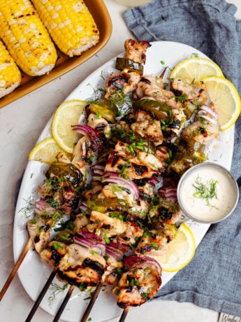 A platter of lemonade pickle brined grilled chicken skewers topped with fresh dill and chives, layered with charred pickle slices and red onion. The juicy grilled chicken is served with lemon wedges, creamy ranch dipping sauce, and grilled corn on the cob for an easy summer barbecue meal.