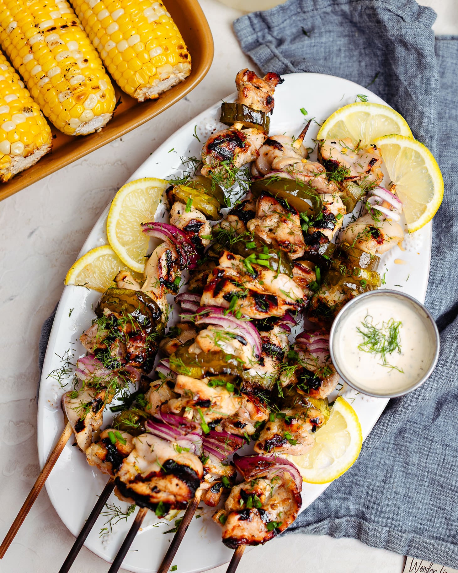 A platter of lemonade pickle brined grilled chicken skewers topped with fresh dill and chives, layered with charred pickle slices and red onion. The juicy grilled chicken is served with lemon wedges, creamy ranch dipping sauce, and grilled corn on the cob for an easy summer barbecue meal.