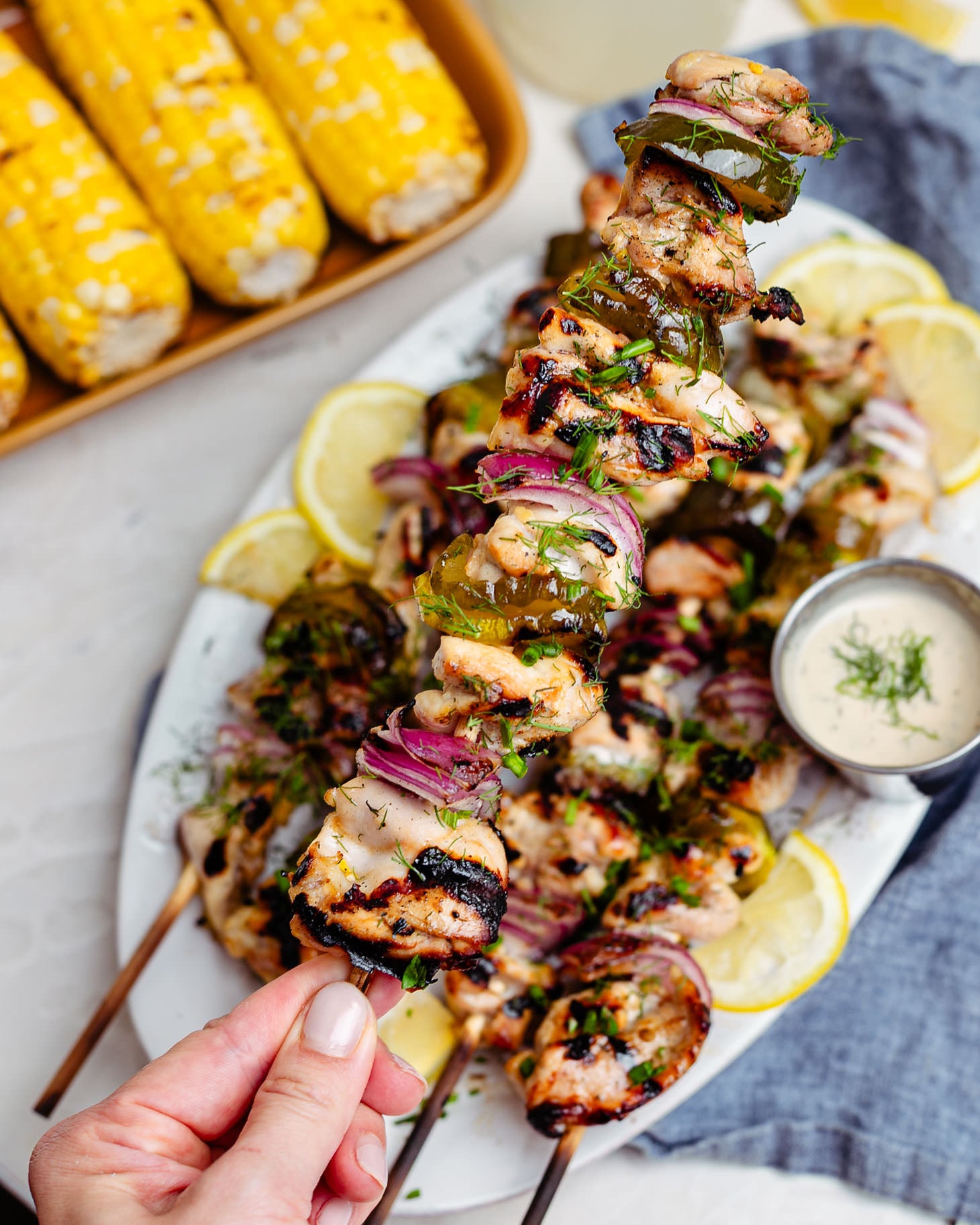 A hand holds up a grilled chicken skewer loaded with juicy charred chicken thighs, dill pickle slices, and red onion. The pickle brined chicken is garnished with fresh dill and chives and served on a platter with lemon slices and a side of creamy ranch dipping sauce. In the background, grilled corn on the cob and a glass of lemonade create the perfect summer cookout scene.