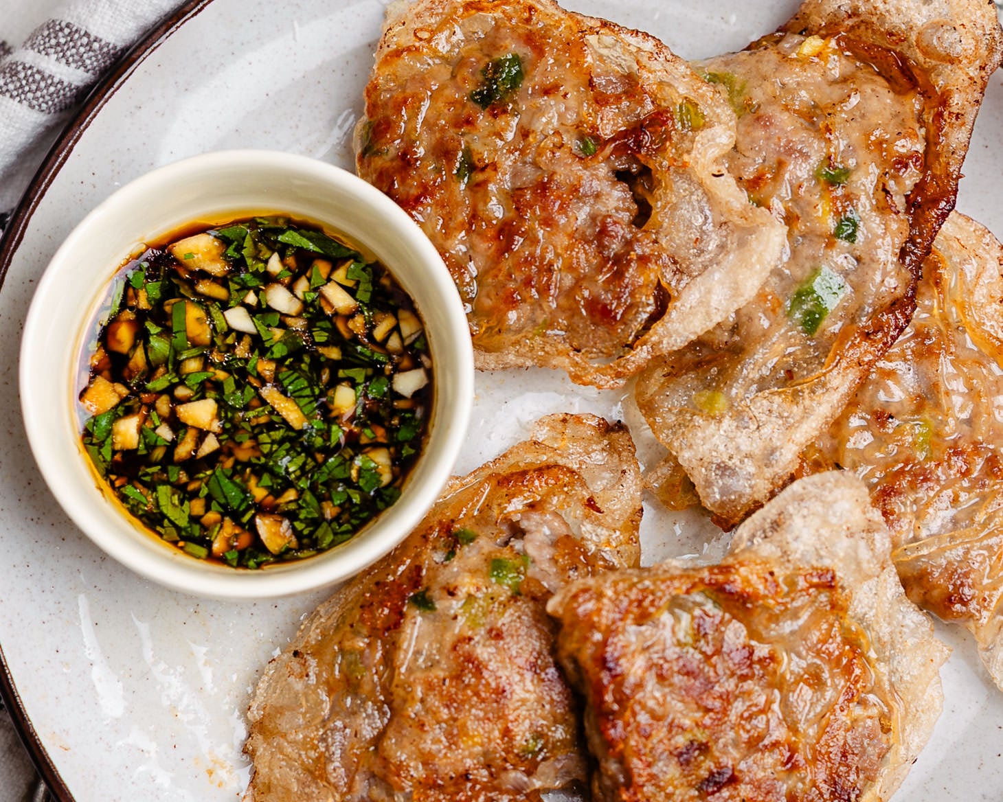 A small dish of dumpling dipping sauce with garlic cilantro soy sauce, black vinegar, sesame oil on a plate along with rice paper dumplings that appear to be crispy golden brown on the outside.