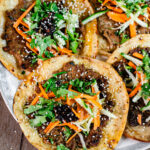 Close-up of a crispy dumpling taco featuring a golden flour tortilla with juicy pork dumpling filling, topped with fresh cucumbers, carrots, cilantro, chili crisp, and sesame seeds, with another taco blurred in the background.
