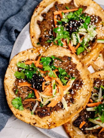 Close-up of a crispy dumpling taco featuring a golden flour tortilla with juicy pork dumpling filling, topped with fresh cucumbers, carrots, cilantro, chili crisp, and sesame seeds, with another taco blurred in the background.