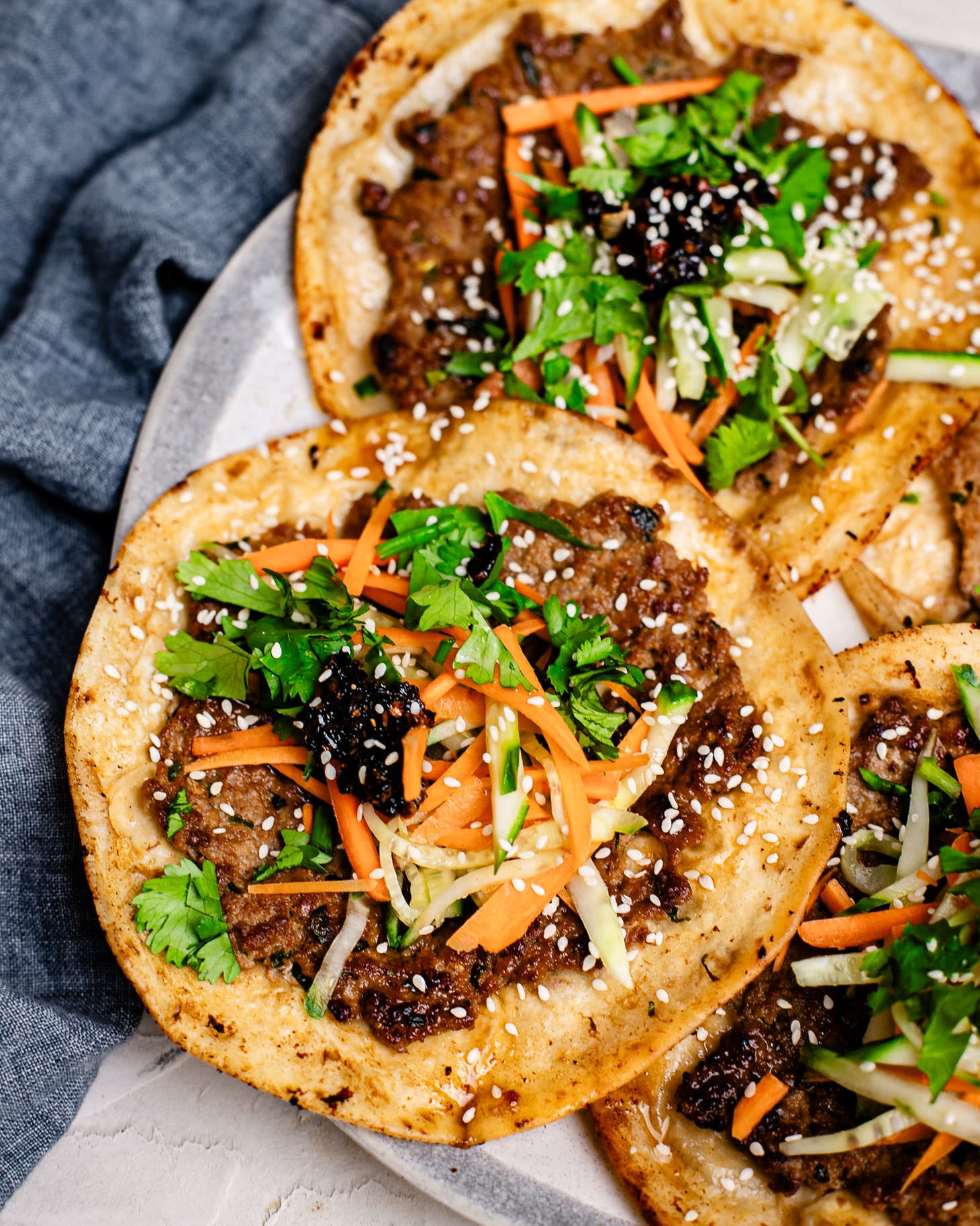 Close-up of a crispy dumpling taco featuring a golden flour tortilla with juicy pork dumpling filling, topped with fresh cucumbers, carrots, cilantro, chili crisp, and sesame seeds, with another taco blurred in the background.