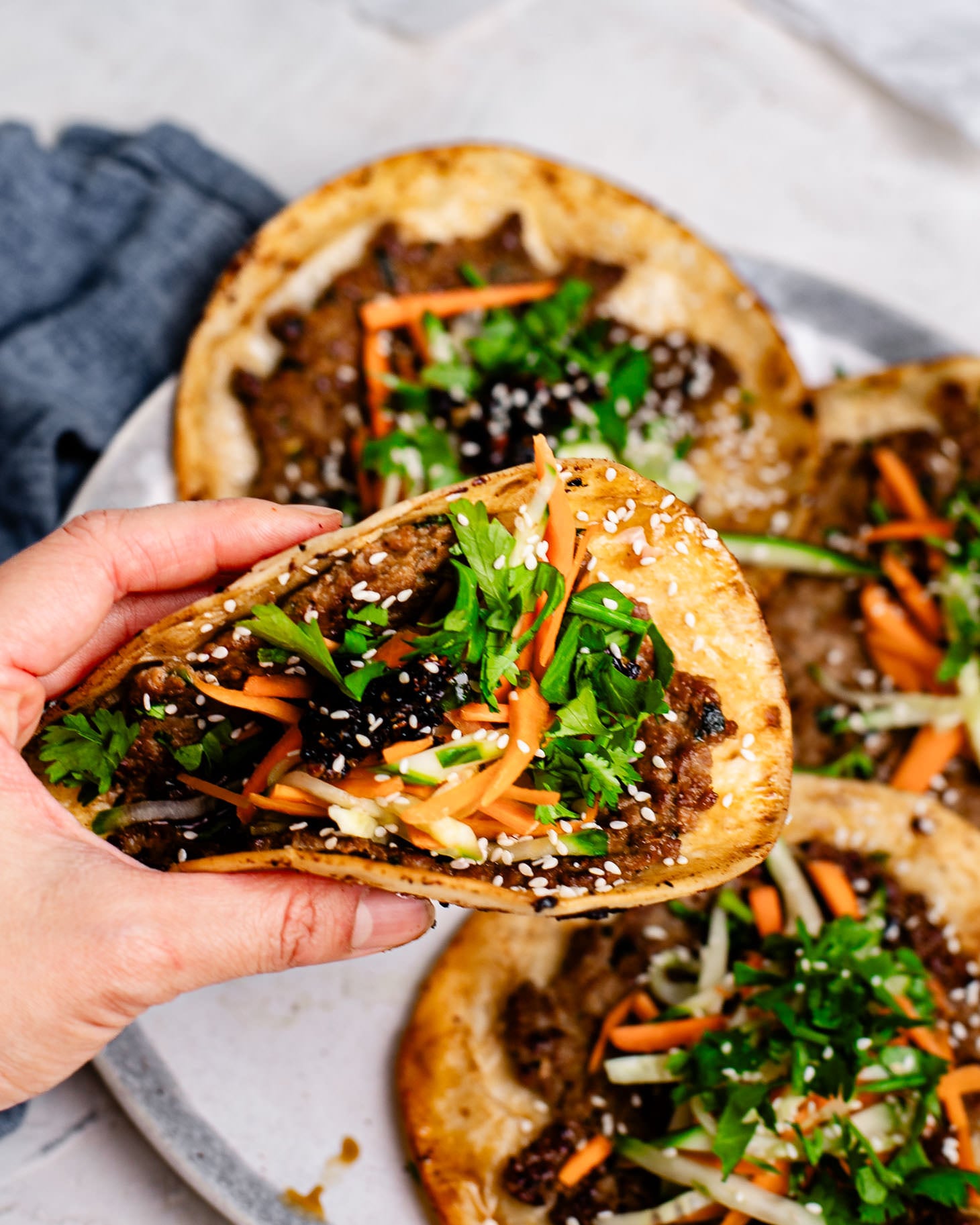 Close-up of a hand holding a golden, crispy dumpling taco filled with pork and topped with fresh carrots, cucumbers, cilantro, chili crisp, and sesame seeds, with a plate of tacos behind it.