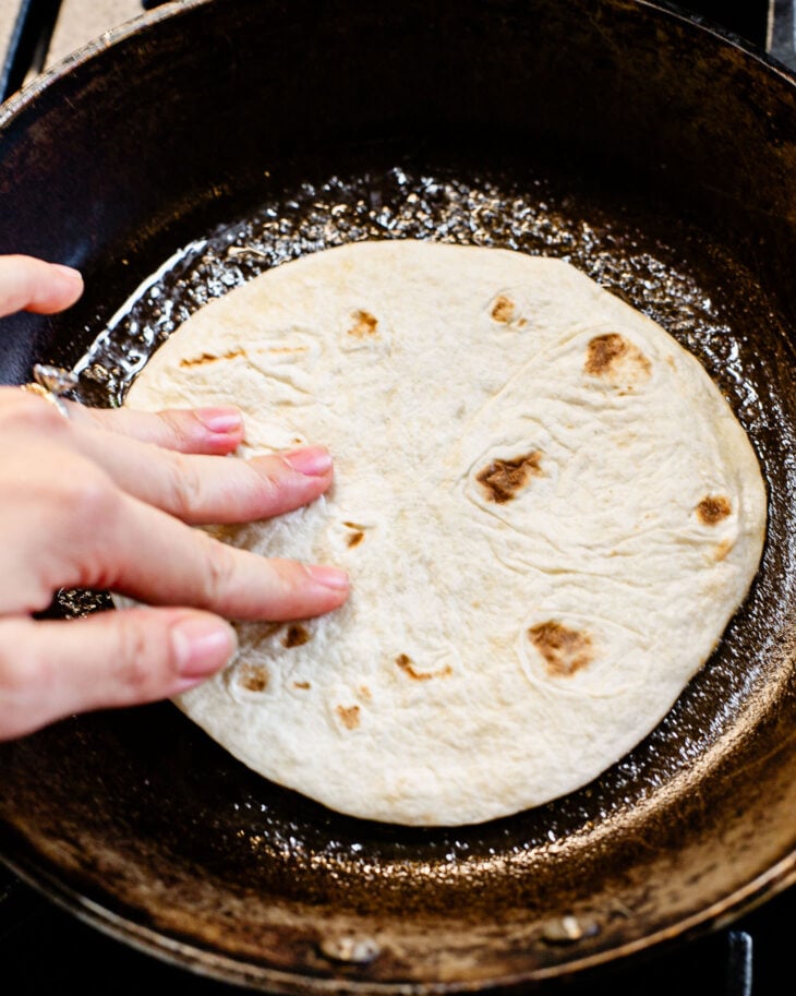 A hand is placing the tortilla filling-side down into the skillet. Gently press with a spatula to spread the meat into an even, thin layer.