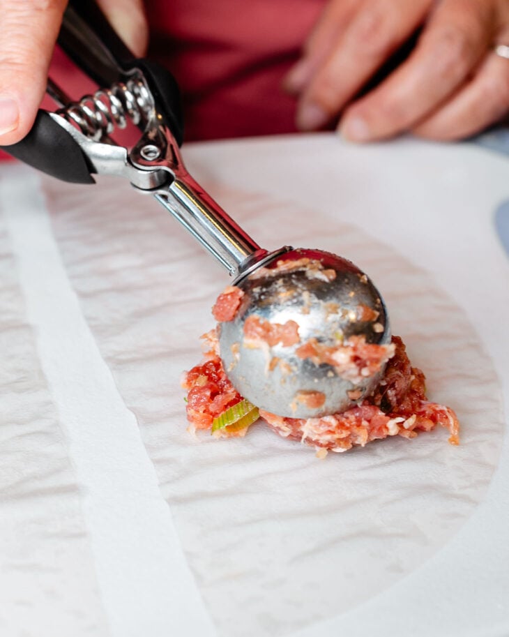 A hand using a scooper to place dumpling filling in the center.