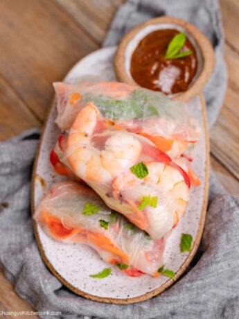 Fresh homemade shrimp spring rolls filled with juicy shrimp, cucumbers, carrots, red bell peppers, and mint, served with creamy peanut dipping sauce on a ceramic platter.