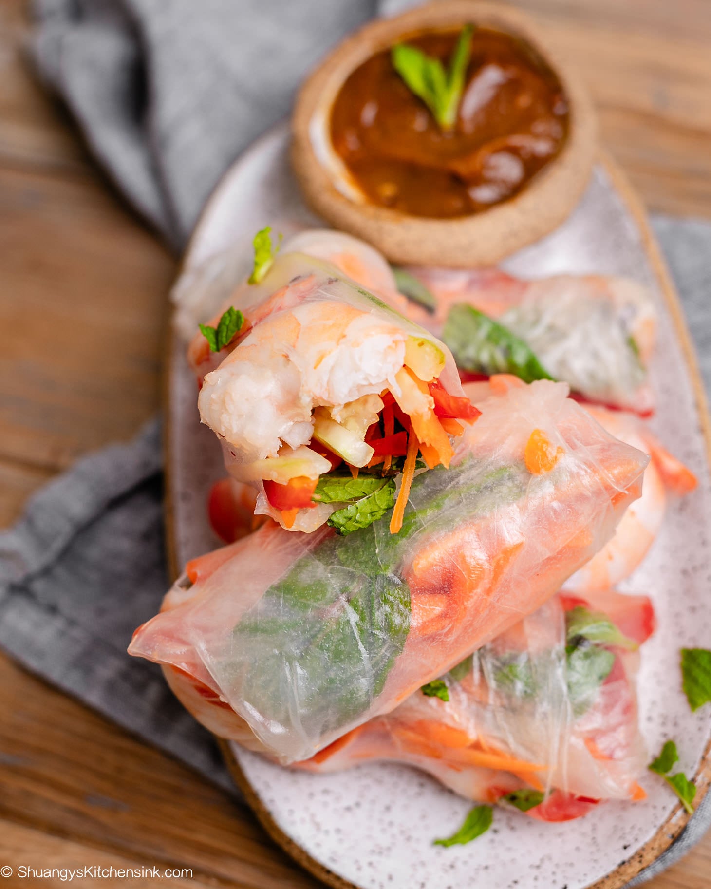 A bite is taken out of a fresh homemade shrimp spring rolls filled with juicy shrimp, cucumbers, carrots, red bell peppers, and mint, served with creamy peanut dipping sauce on a ceramic platter.