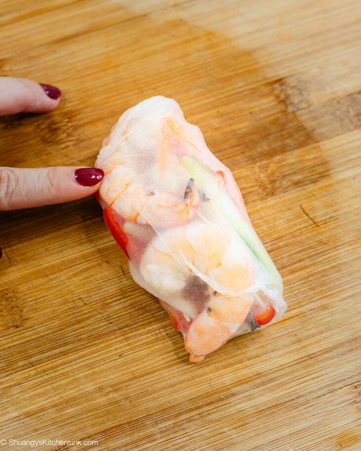 A finger touching a finished spring roll with shrimp, cucumber, red pepper wrapped in it.