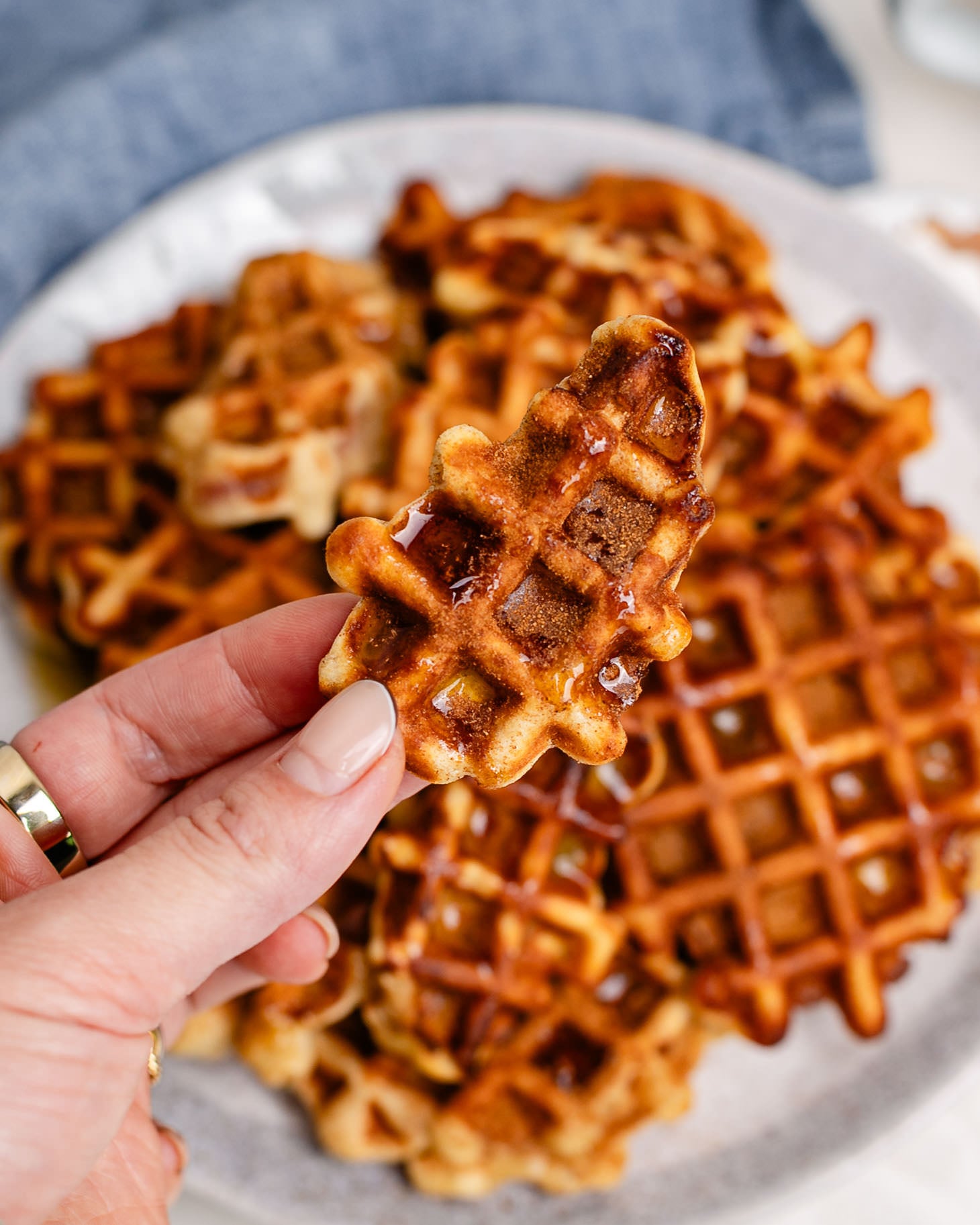 Apple pie waffle bite topped with cinnamon sugar and maple syrup, held over a plate of golden waffle bites.